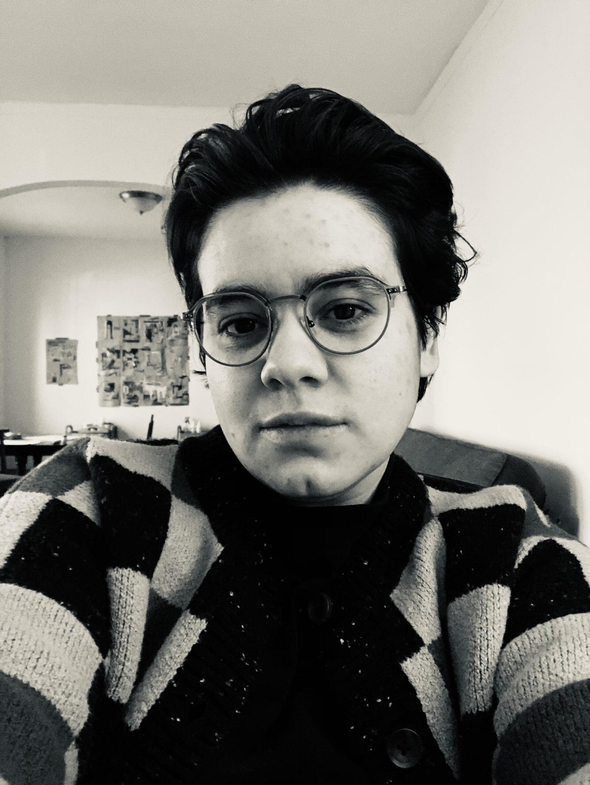 A black and white photo shows Ariana, a white, Latinx, nonbinary person with short, dark hair wearing round eyeglasses and a checkerboard patterned sweater. Collages, magazine clippings, and other art studio ephemera hang on the wall behind them.