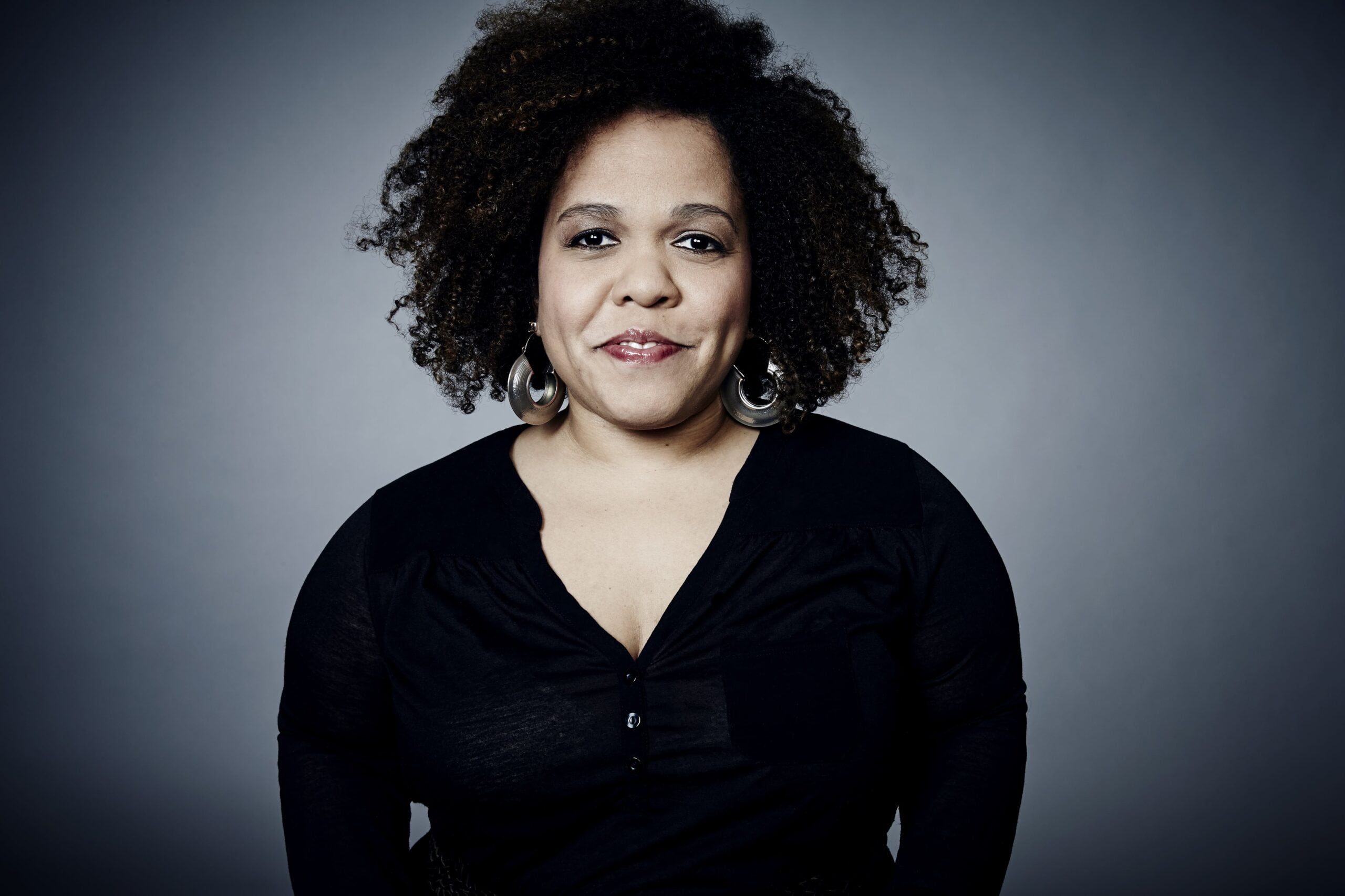a light skin Black woman with big curly hair looks directly in the camera smiling wearing a Black v neck shirt and big silver hoops.