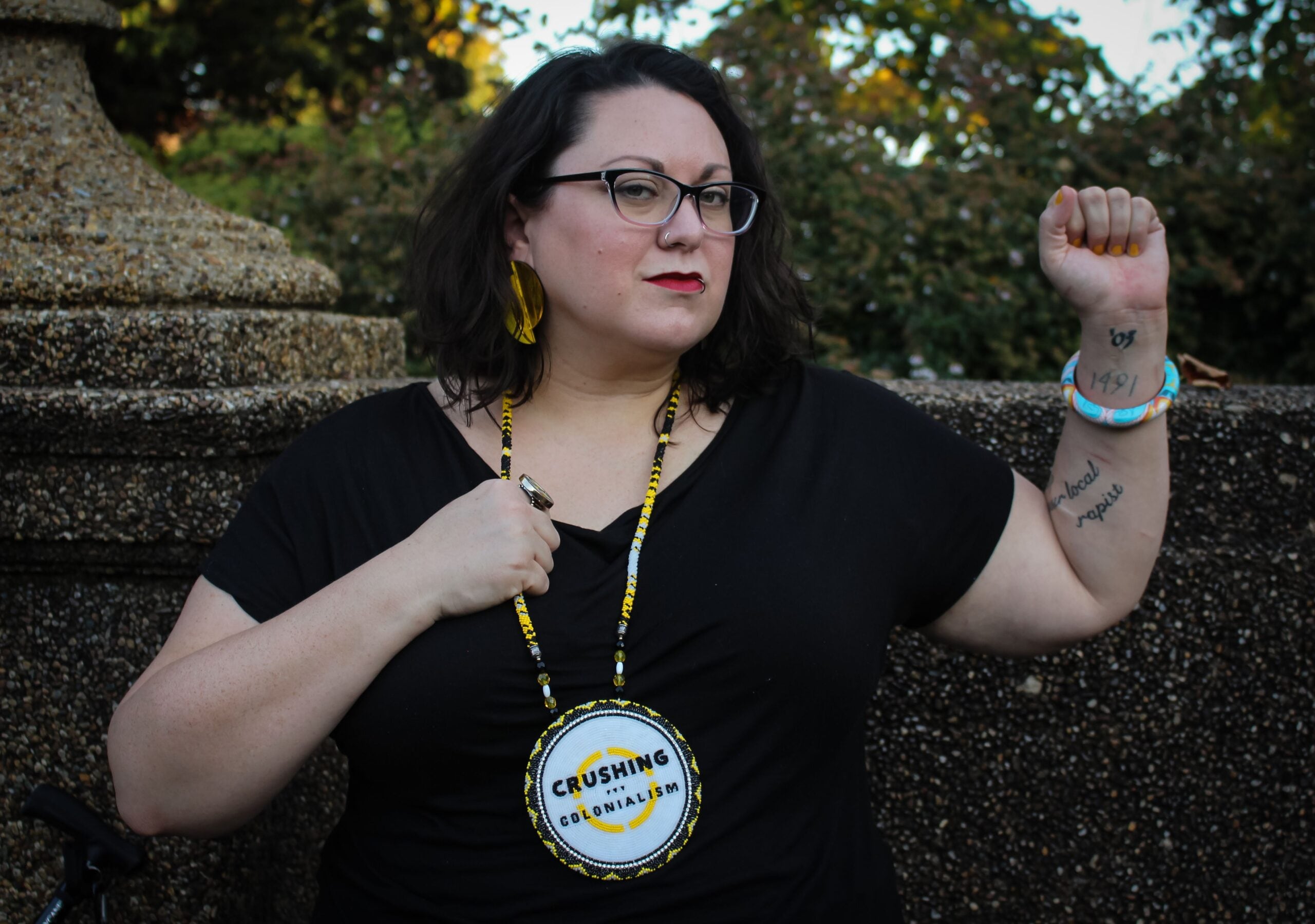 Jen is wearing a black tee, their yellow, white and black beaded medallion with Crushing Colonialism logo, black glasses, their brown hair is parted to the side and worn down and wavy, and large yellow earrings and light blue bracelet on their fist raised in the air.