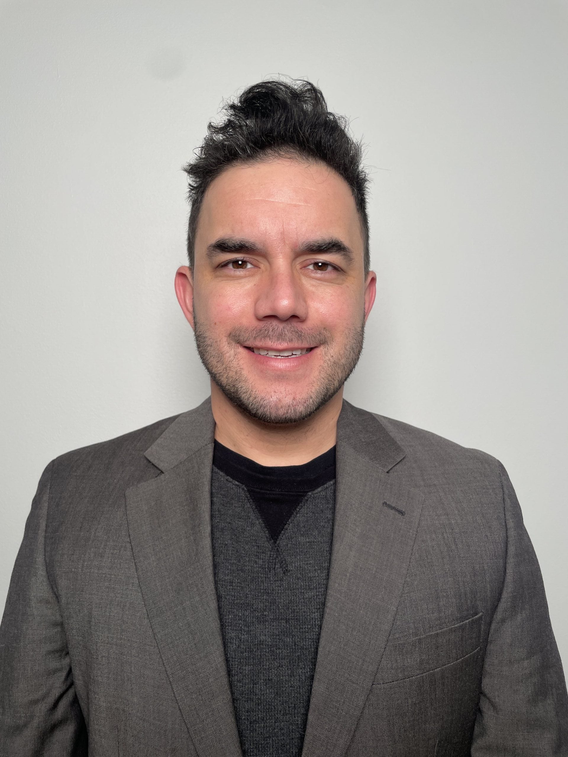 A Hispanic man with curly brown hair and brown eyes wearing a grey blazer looks into the camera.