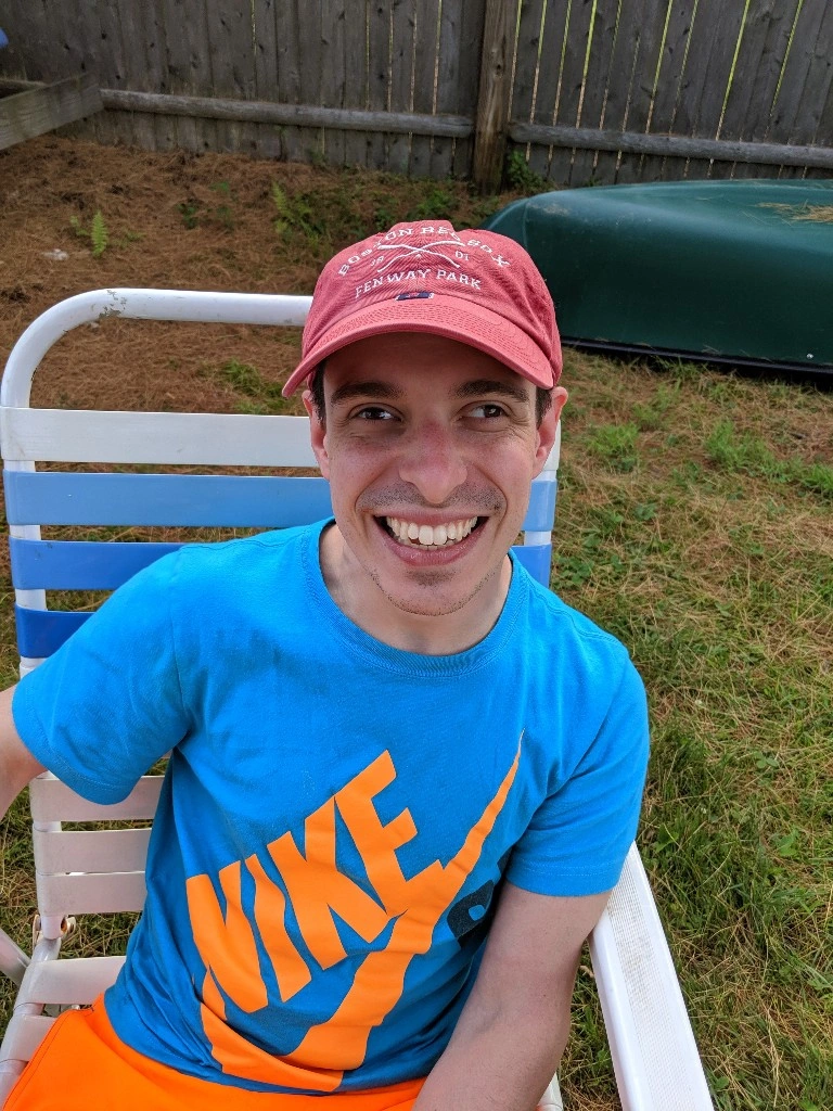 A man in a blue Nike shirt with yellow writing and a red baseball cap looks directly into the camera while sitting on a lawn chair.