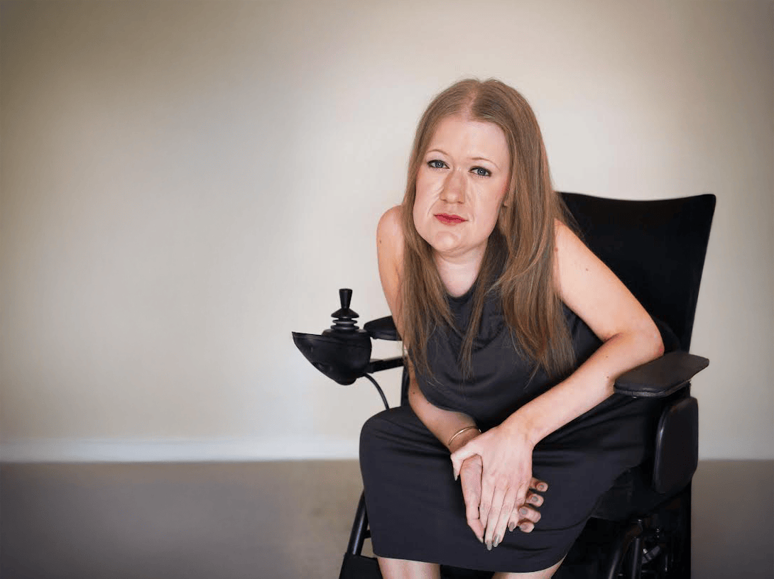a white woman with blonde hair sat in a wheelchair. I’m wearing a sleeveless brown/taupe dress.