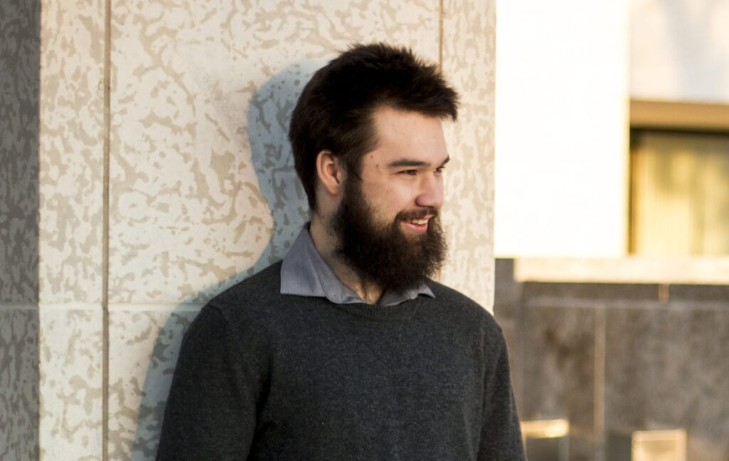 John, a white man with a beard, is standing in front of a column with his hands in his pockets.