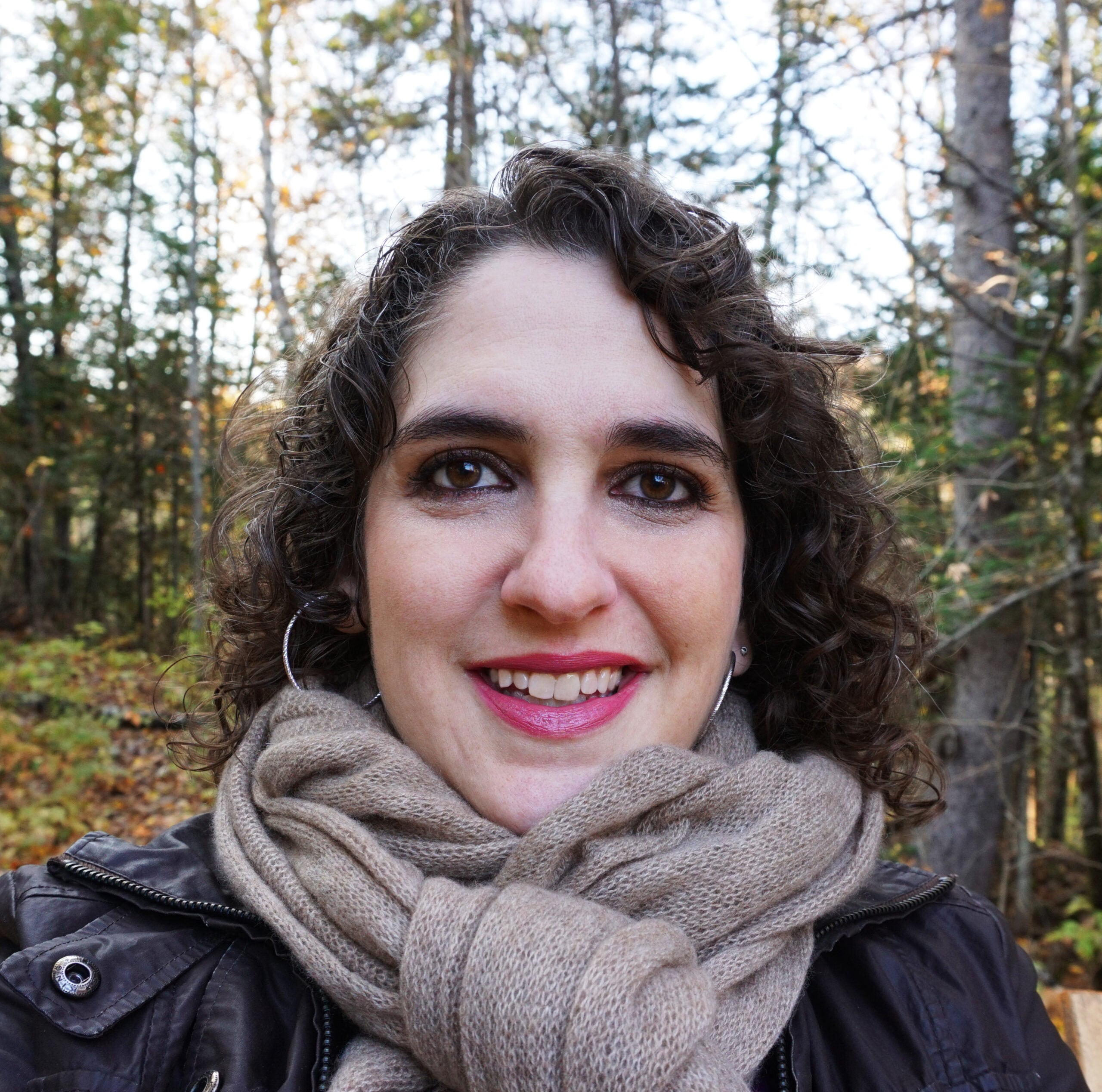 A white woman with shoulder-length curly brown hair wearing large silver hoop earrings and a tan scarf bundled around her neck, with an autumn forest landscape behind her.