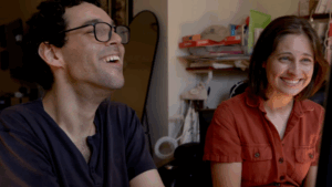Director Reid Davenport, a white man with curly, short brown hair and glasses, and Producer Colleen Cassingham, a white woman with a brown bob wearing an orange jumpsuit, laugh together as they look toward a computer screen, which is just out of the shot. In the background, is an apartment with a full length mirror and an array of books on a shelf.