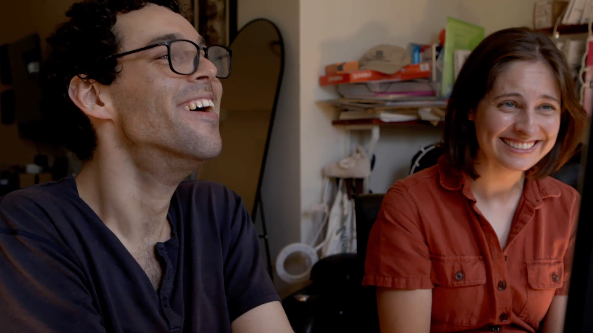 Director Reid Davenport, a white man with curly, short brown hair and glasses, and Producer Colleen Cassingham, a white woman with a brown bob wearing an orange jumpsuit, laugh together as they look toward a computer screen, which is just out of the shot. In the background, is an apartment with a full length mirror and an array of books on a shelf.