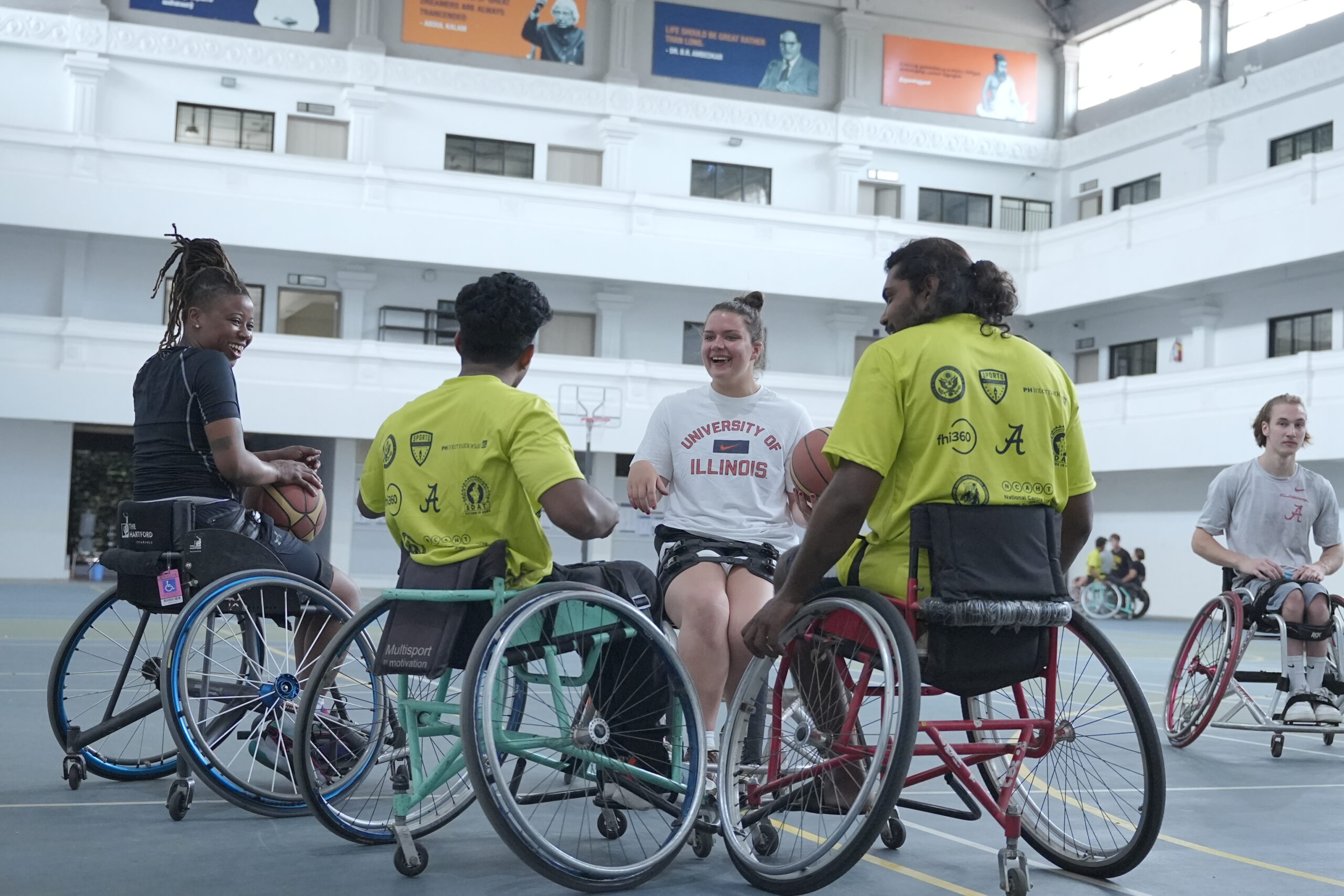 A group of four wheelchair basketball players gathered on a court laughing with one another.
