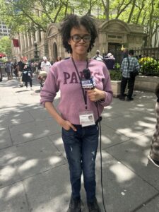 Ashley Castillo, a Black Afro-Latina journalist, stands in a pink shirt and jeans. She is holding a TV microphone and is smiling. Behind her is a street scene that includes people and trees
