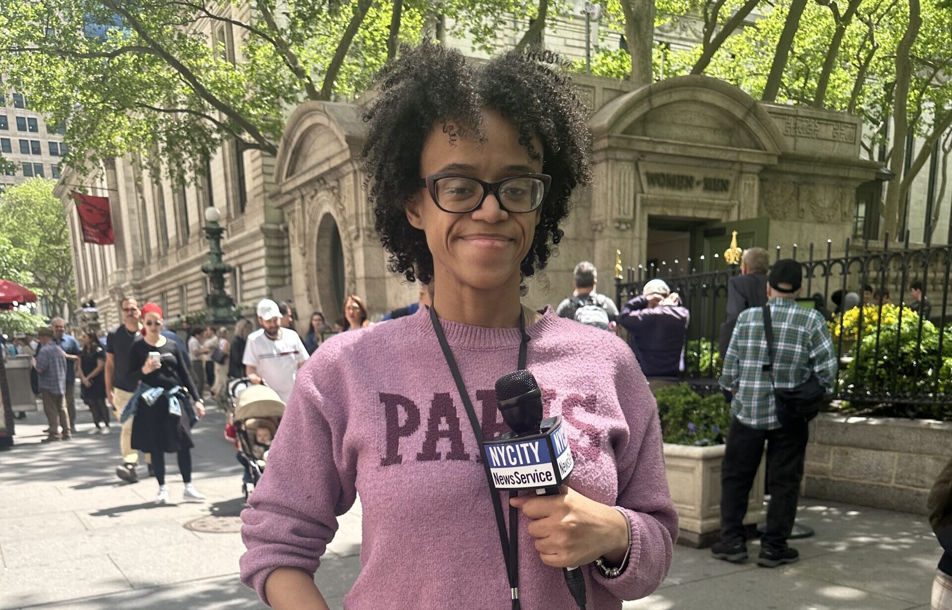 Ashley Castillo, a Black Afro-Latina journalist, stands in a pink shirt and jeans. She is holding a TV microphone and is smiling. Behind her is a street scene that includes people and trees