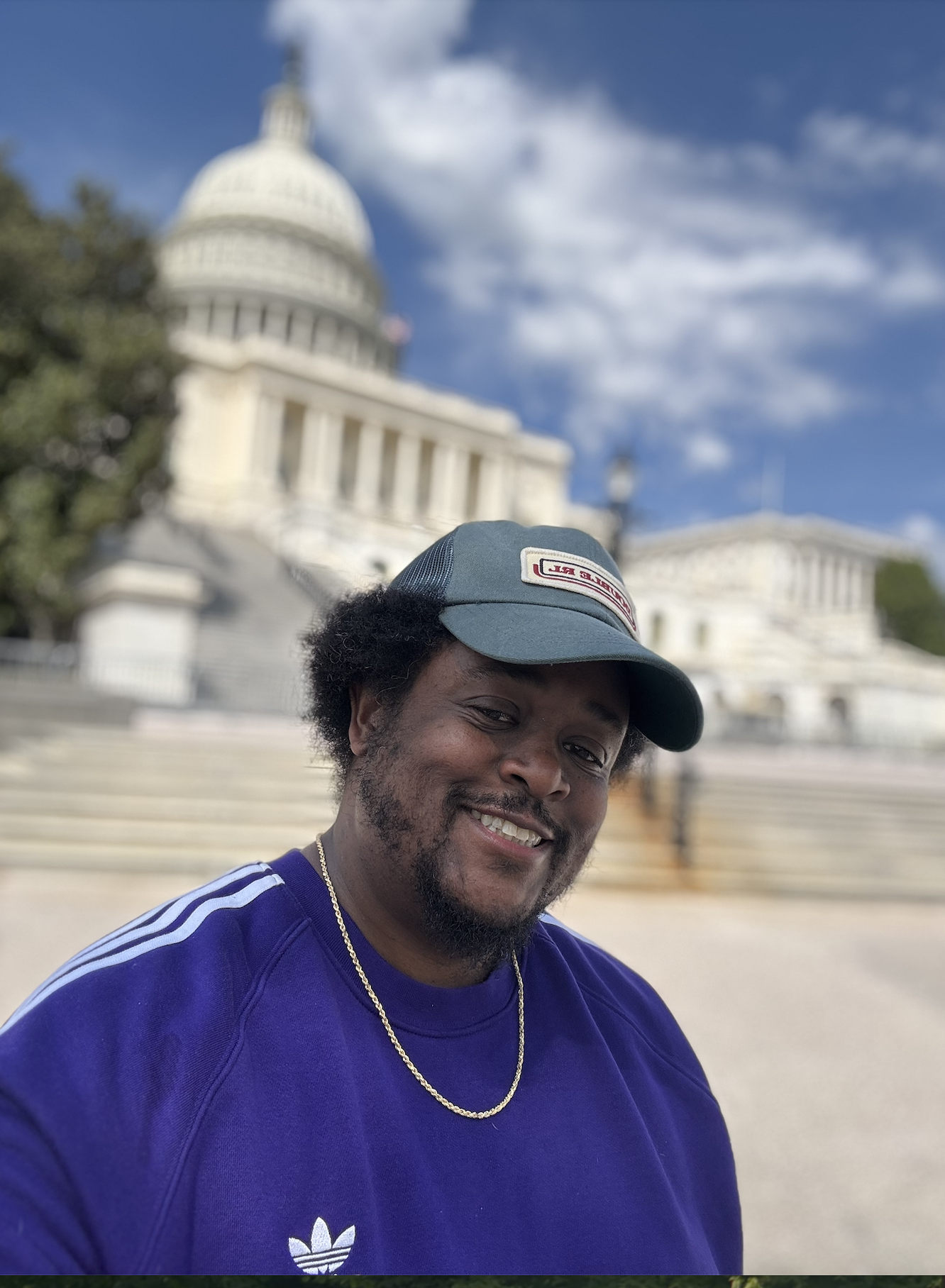 Ryan, a black man in a purple shirt and blue hat, is smiling in front of the steps of a white legislative building.