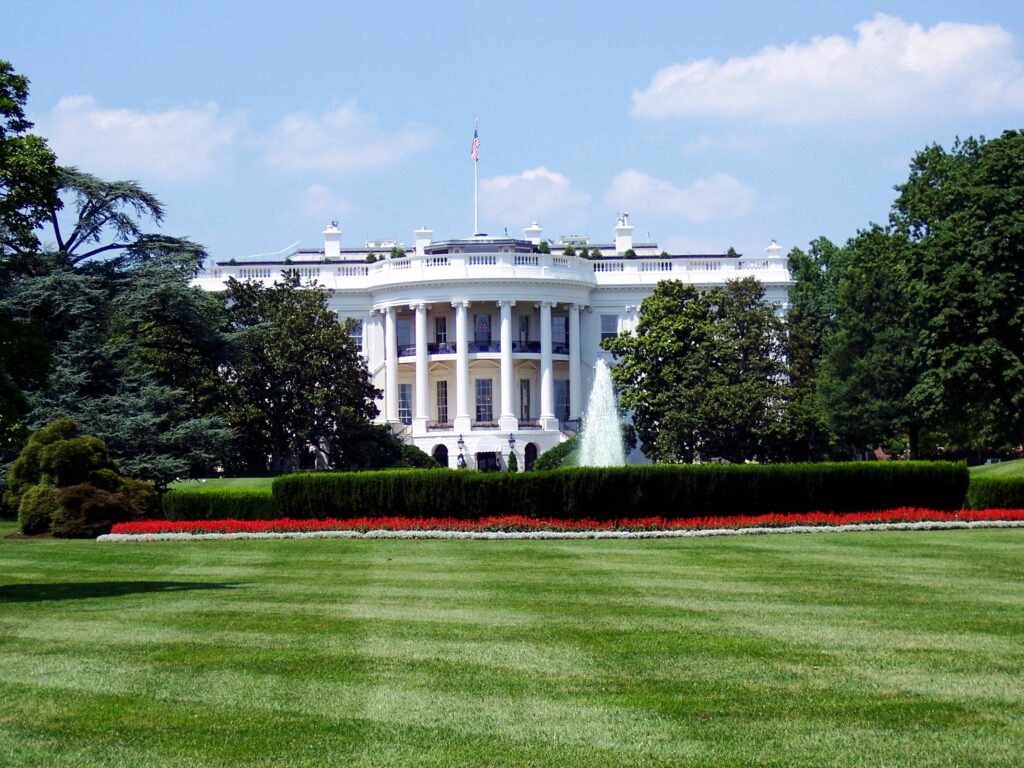 An image of the White House, fountain in front in full view. There are a few clouds above in a blue sky