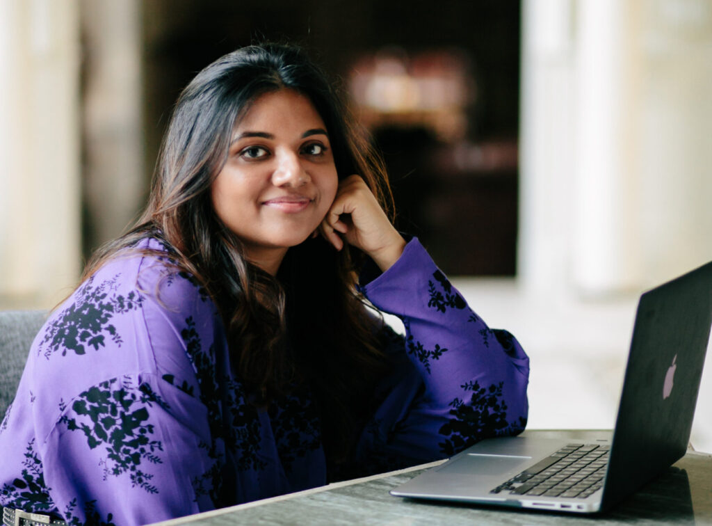 Journalist Sonali Gupta wearing a purple shirt with black pint, seated at a table with a laptop open in front of her