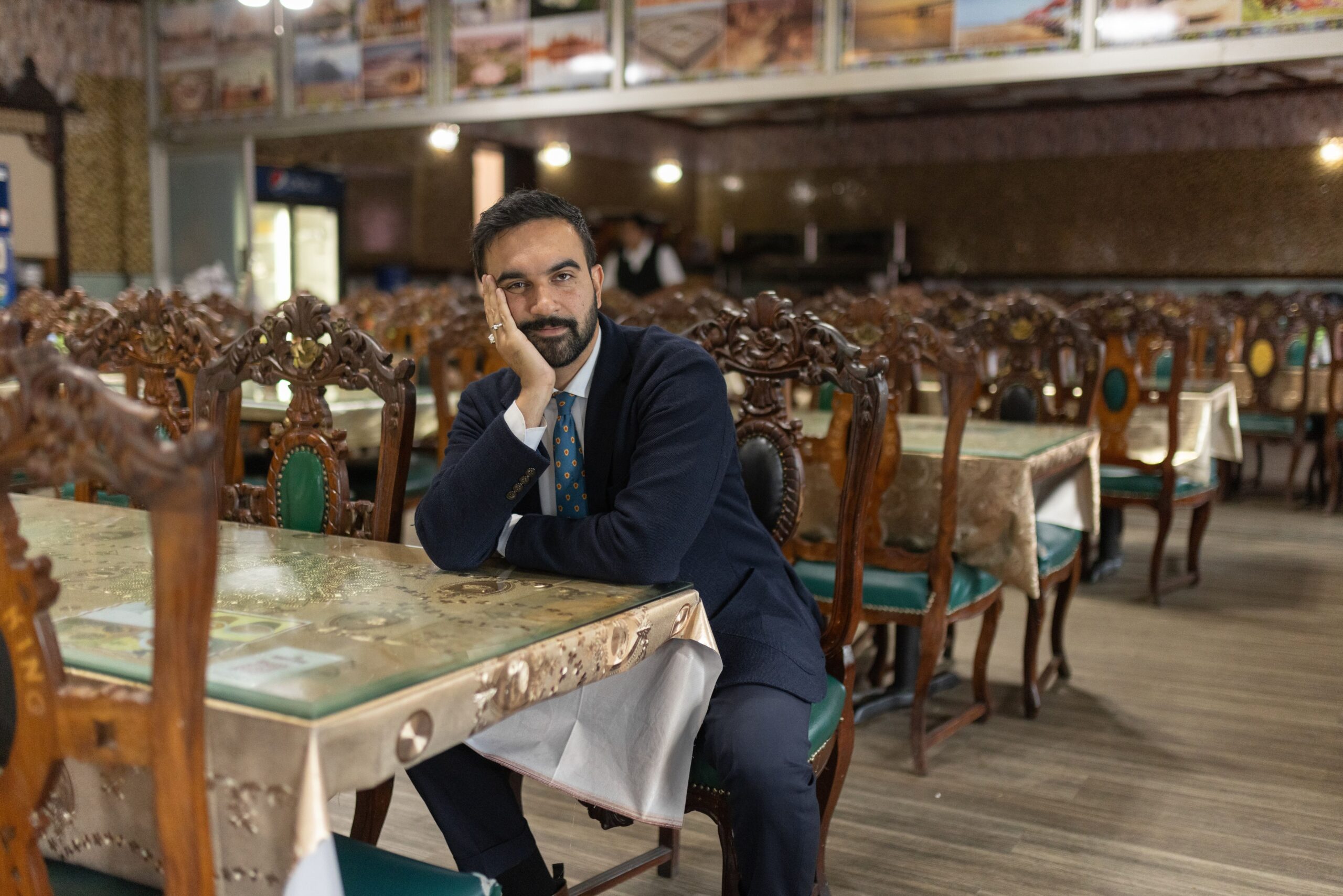 An image of Zohran Mamdani, in a suit, head on his hand, in an empty dining room