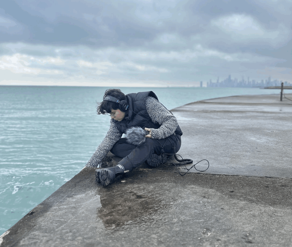 DJA team member Ariana Martinez sits on a rocky bank next to a body of water holding audio recording equipment