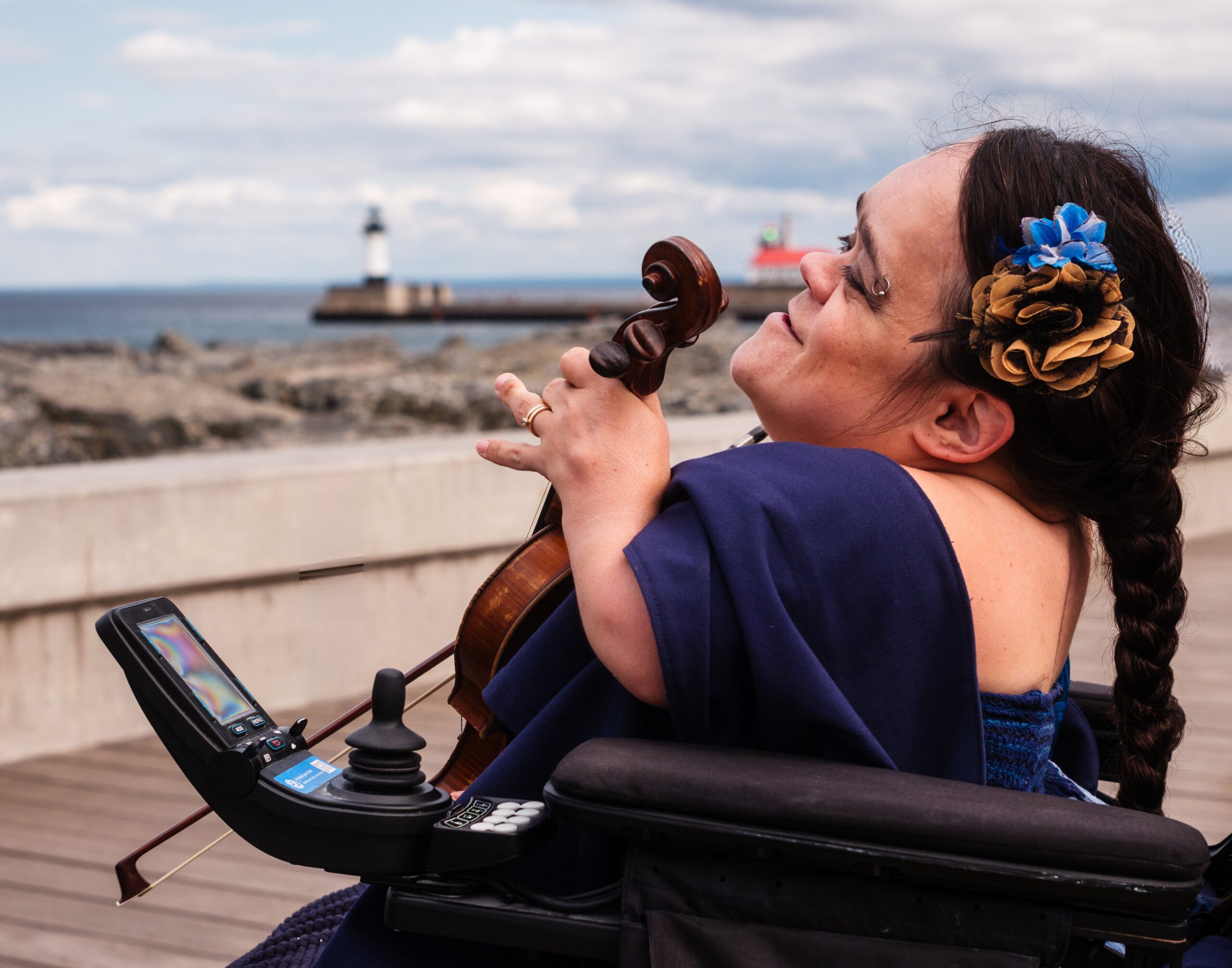 Gaelynn Lea, a white musician, sits in her power wheelchair with her musician. She looking out on a boardwalk