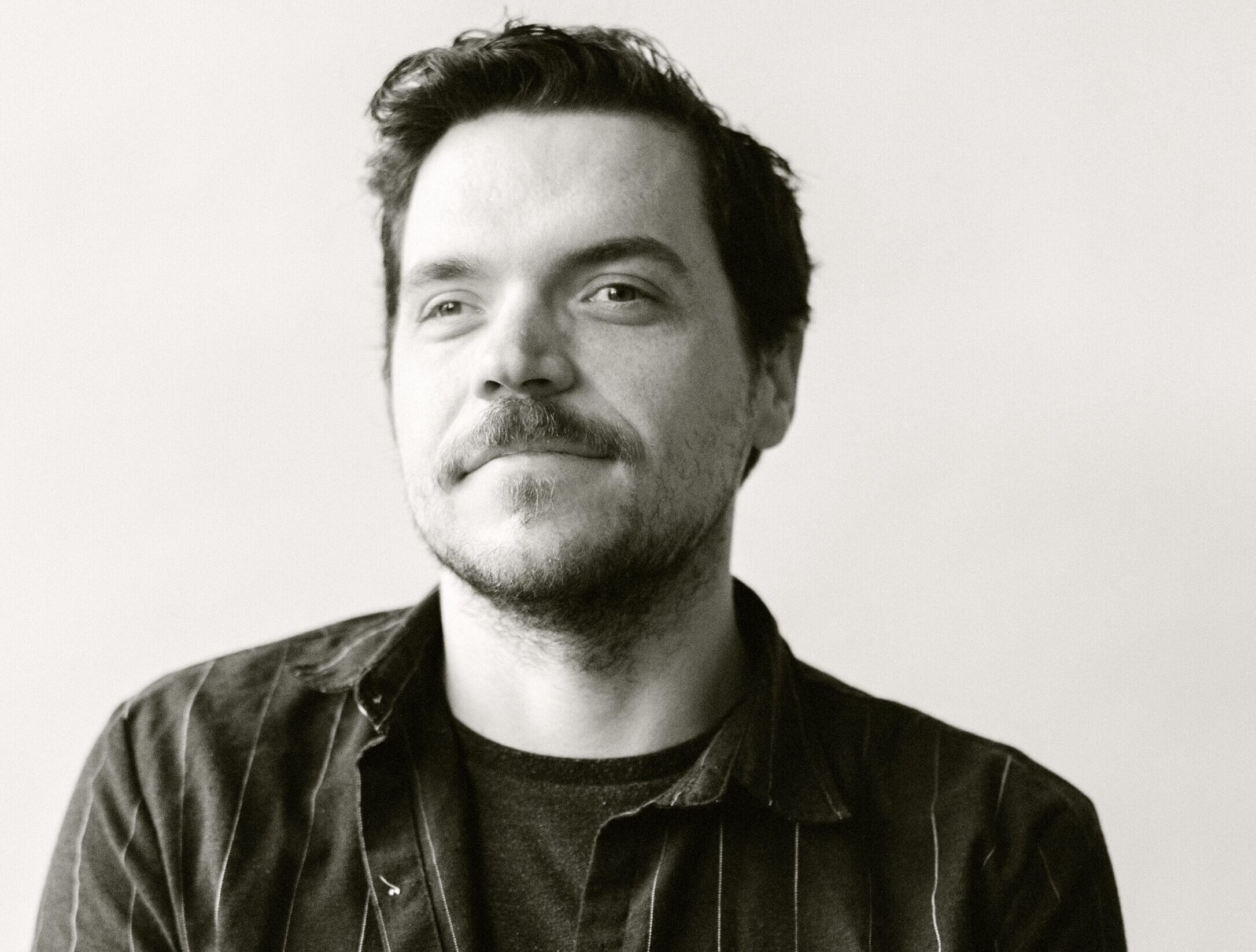 Sean, a white man, stands against a white background with a half buttoned pin stripe shirt. The photo is in black and white.