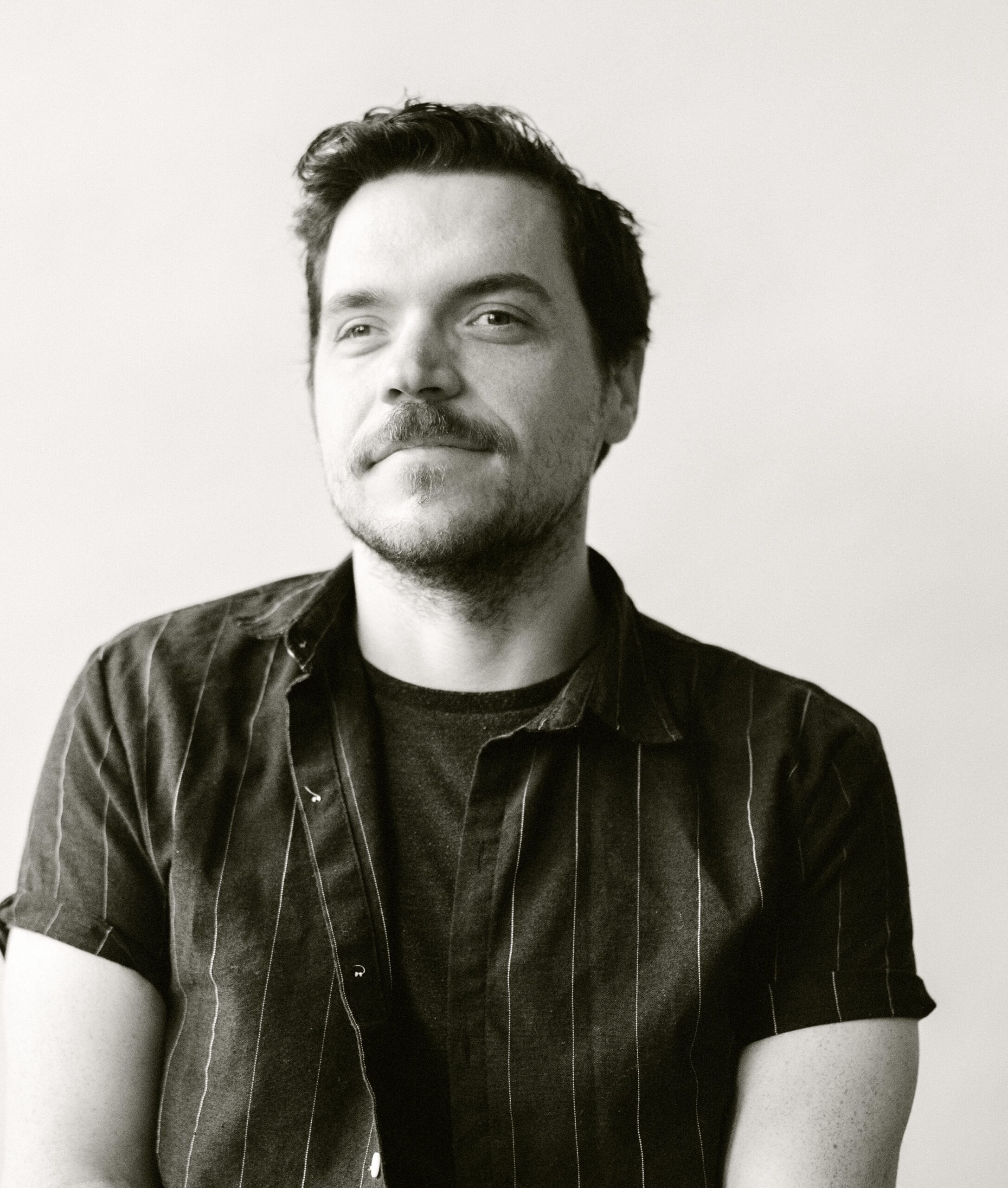 sk headshot 2025 Sean, a white man, stands against a white background with a half buttoned pin stripe shirt. The photo is in black and white.