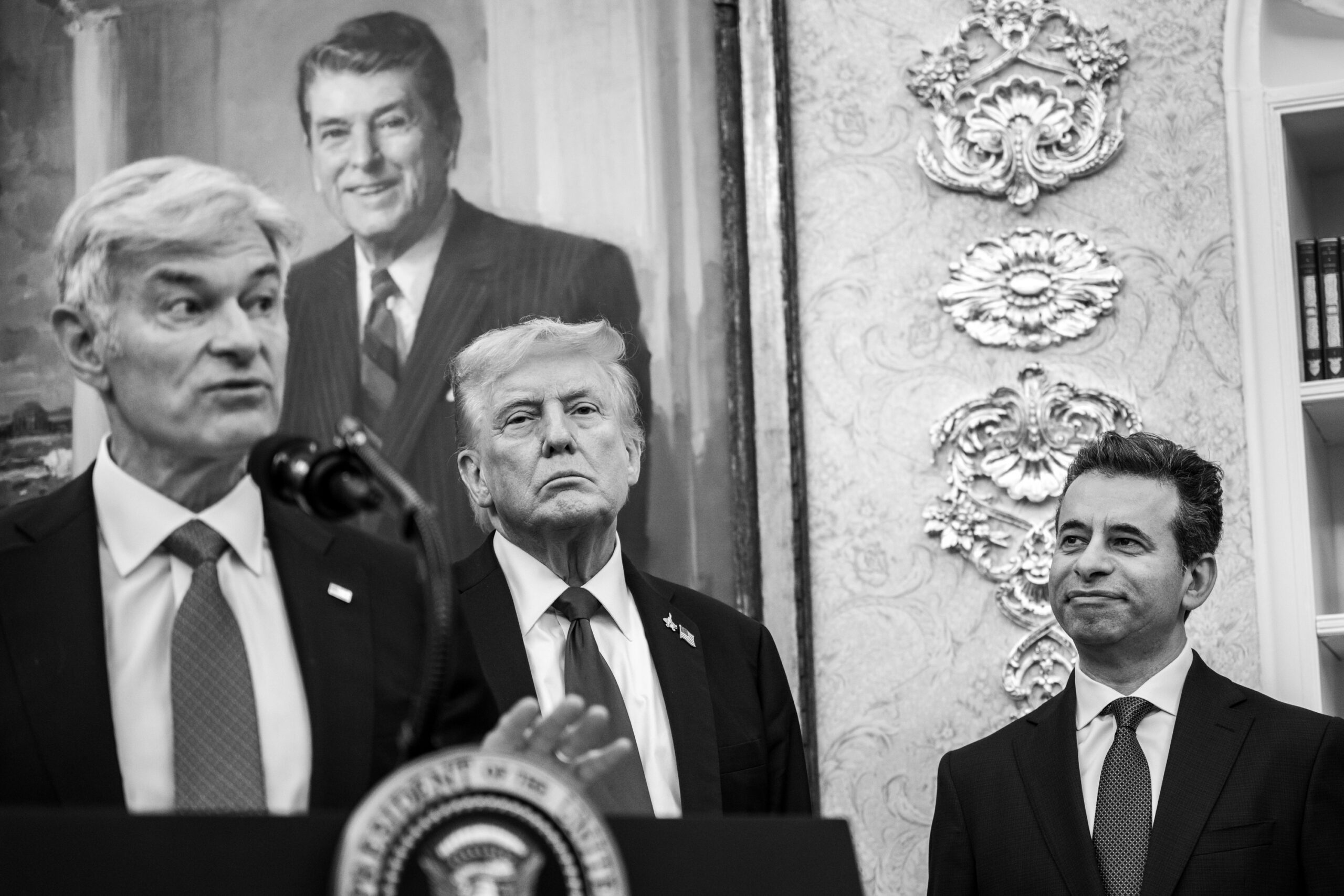 Black-and-white photograph of CMS Administrator Dr. Mehmet Oz speaking at a podium in the Oval Office, gesturing as he delivers remarks. President Donald Trump stands behind him at center with a serious expression, while another man in a suit stands to the right, watching with a slight smile. The background features ornate wall detailing and a large portrait of former President Ronald Reagan.
