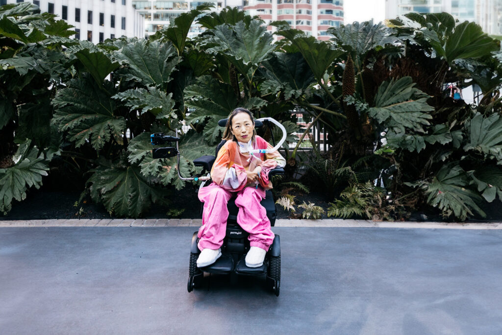 Alice Wong sits in a motorized wheelchair on a paved path, wearing bright pink pants and a colorful top. A ventilator tube is positioned near her face. She is centered in front of large, lush green plants, with tall buildings visible in the background, creating a contrast between nature and an urban setting.