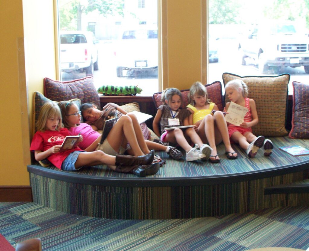 Six children sit together on a curved bench in a bright library space, each reading or looking at books near large windows.