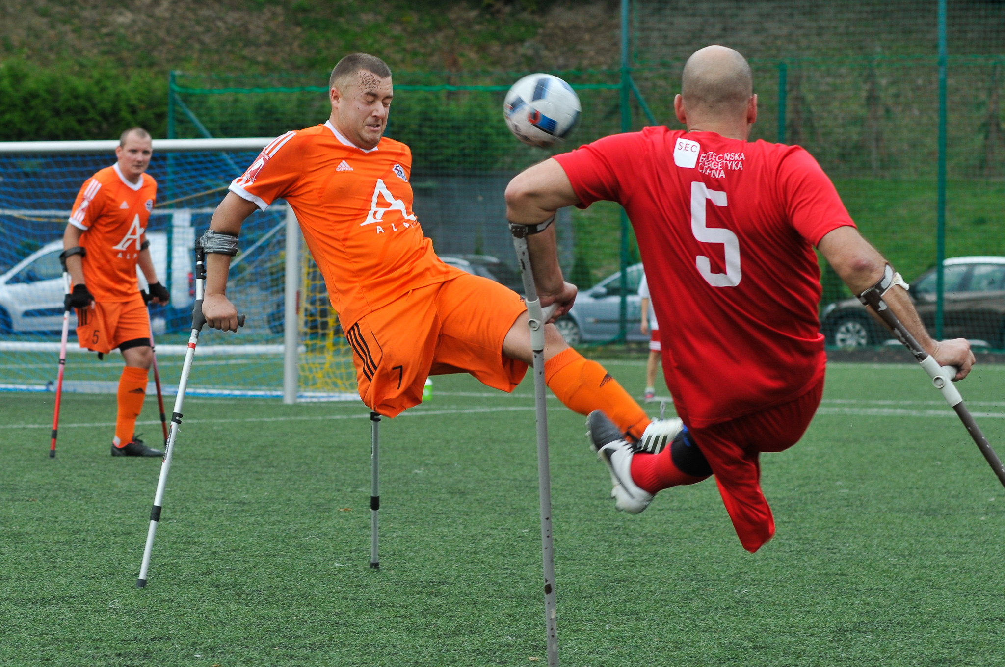 Amputee soccer players compete on a turf field, using crutches as an orange-clad player swings his leg to strike the ball while a defender in red challenges, with teammates and a goal visible in the background.