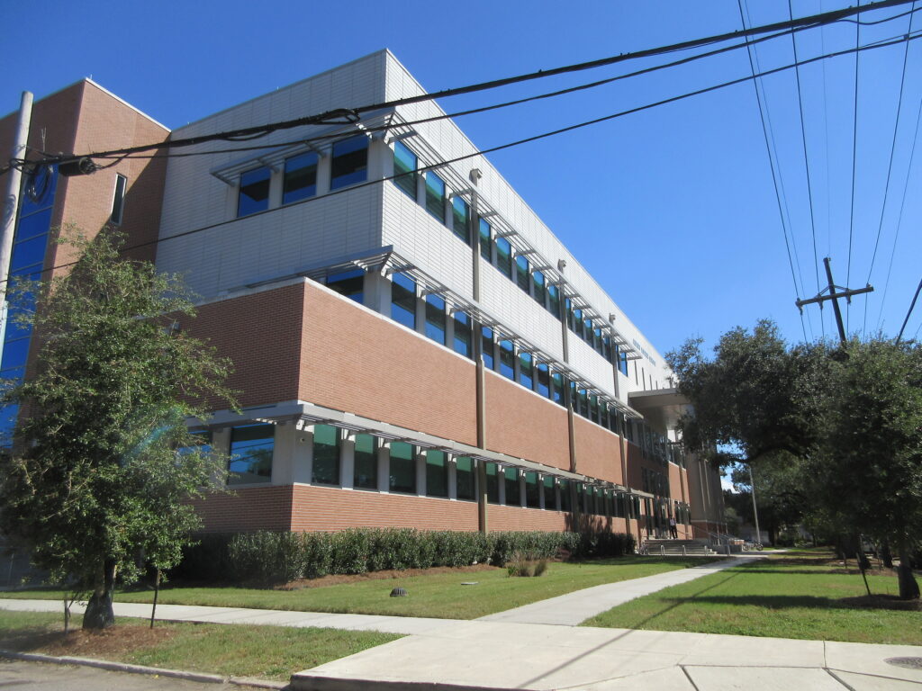 Modern three-story school building with red brick and white panel exterior, rows of large windows, and a covered entrance, seen from a sidewalk on a clear sunny day with trees and power lines in view.