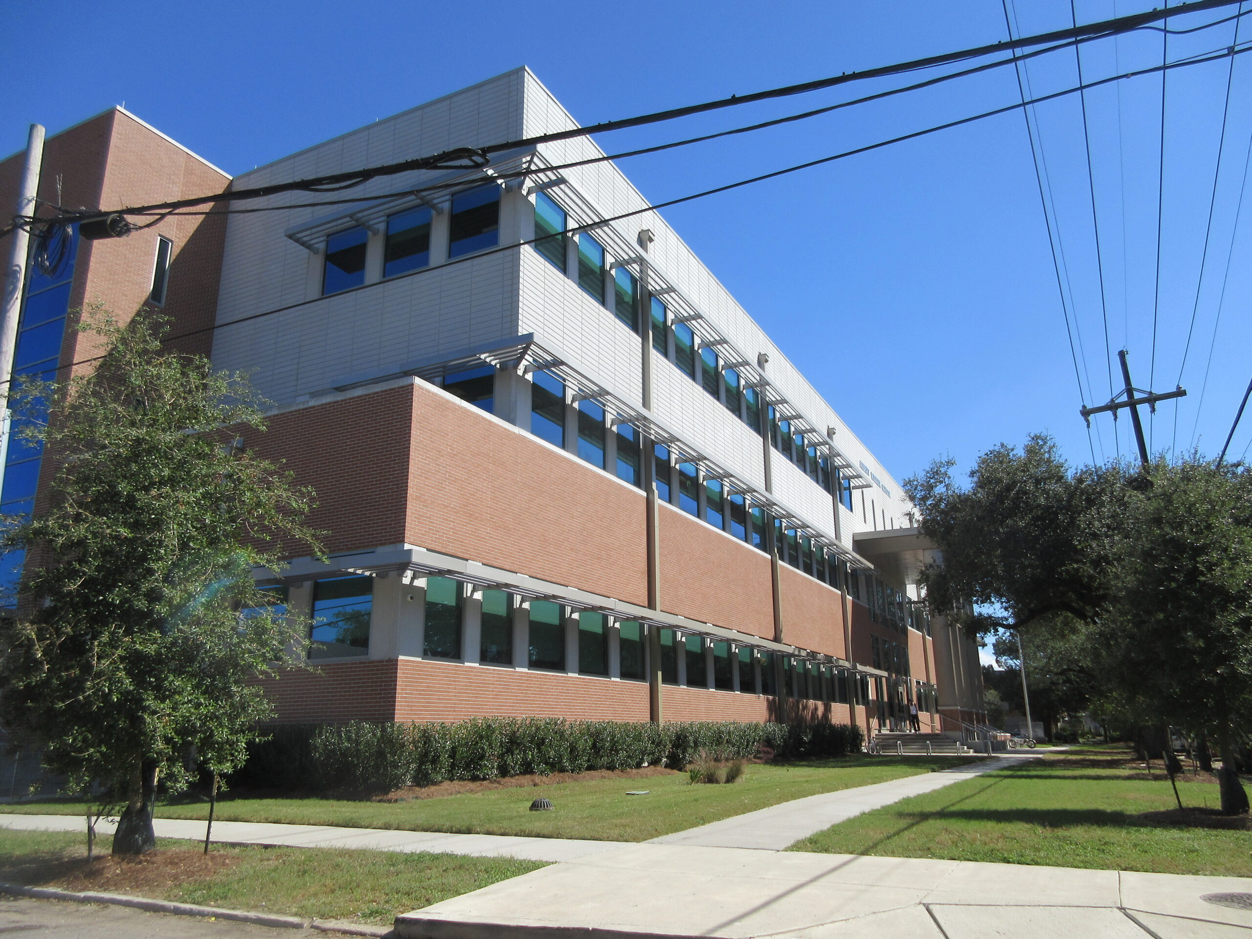 Modern three-story school building with red brick and white panel exterior, rows of large windows, and a covered entrance, seen from a sidewalk on a clear sunny day with trees and power lines in view.