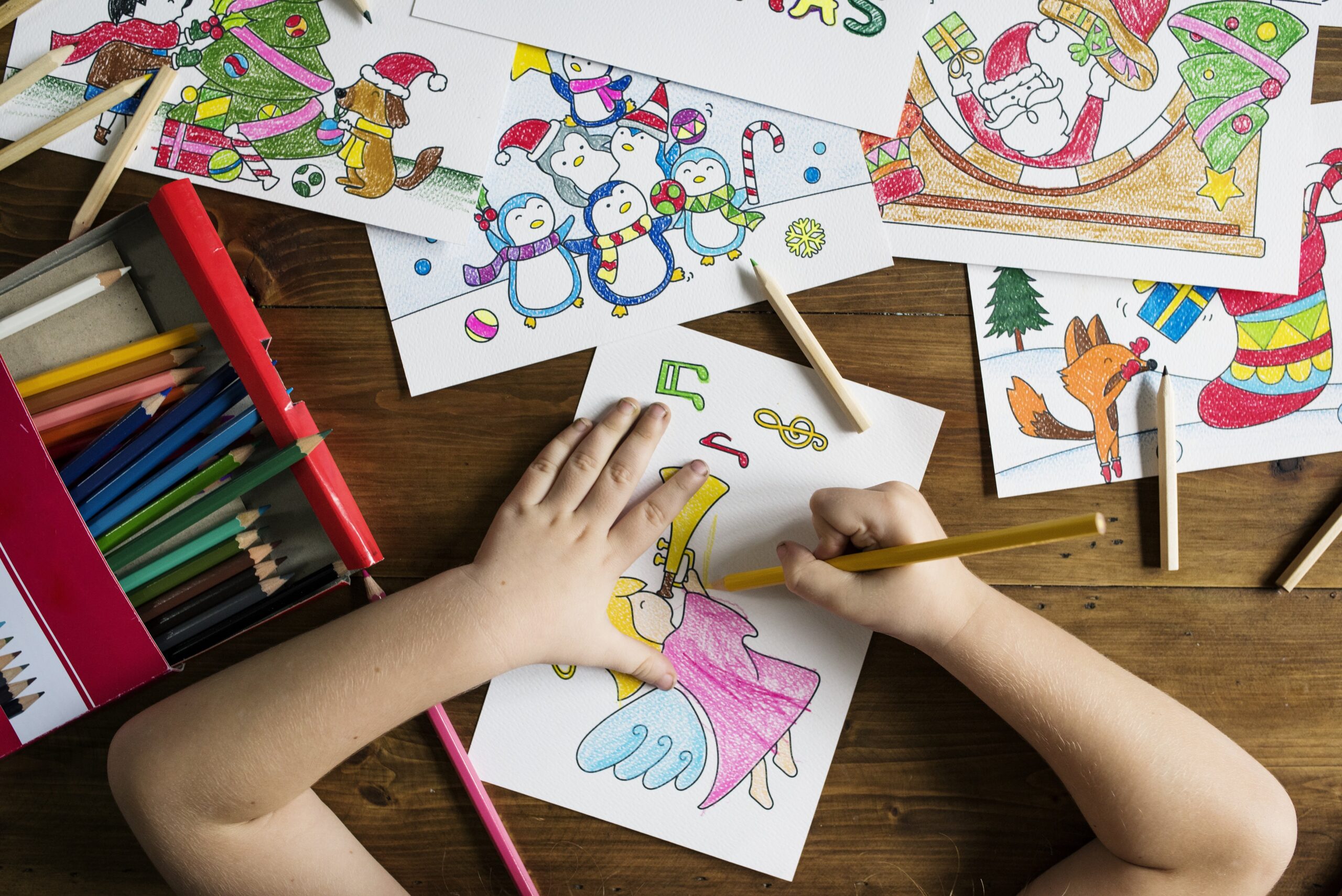 A child drawing at a wooden table. Drawings and pens are seen all over the table.