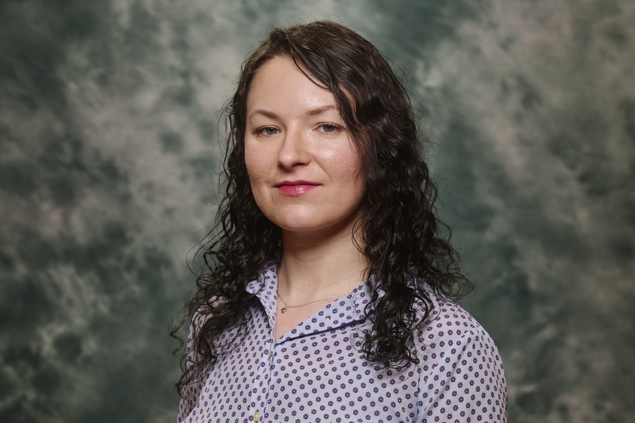 Caroline is wearing a light blue button down shirt with small purple daisies and a necklace with a small blue stone. They have mid length curly brown hair, fair skin, blue eyes, and a calm expression. The background is green and gray marbled.