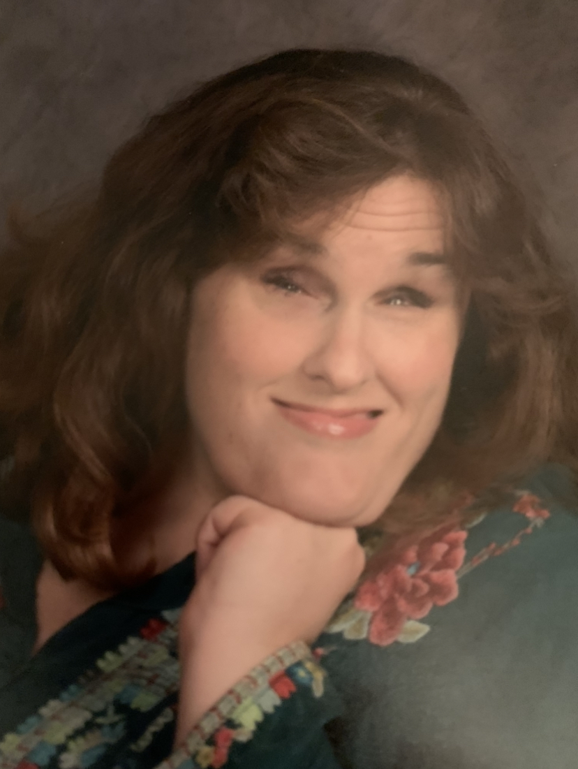 Kelli, sits for a portrait. There is a brown background, she is wearing a green and red floral shirt.