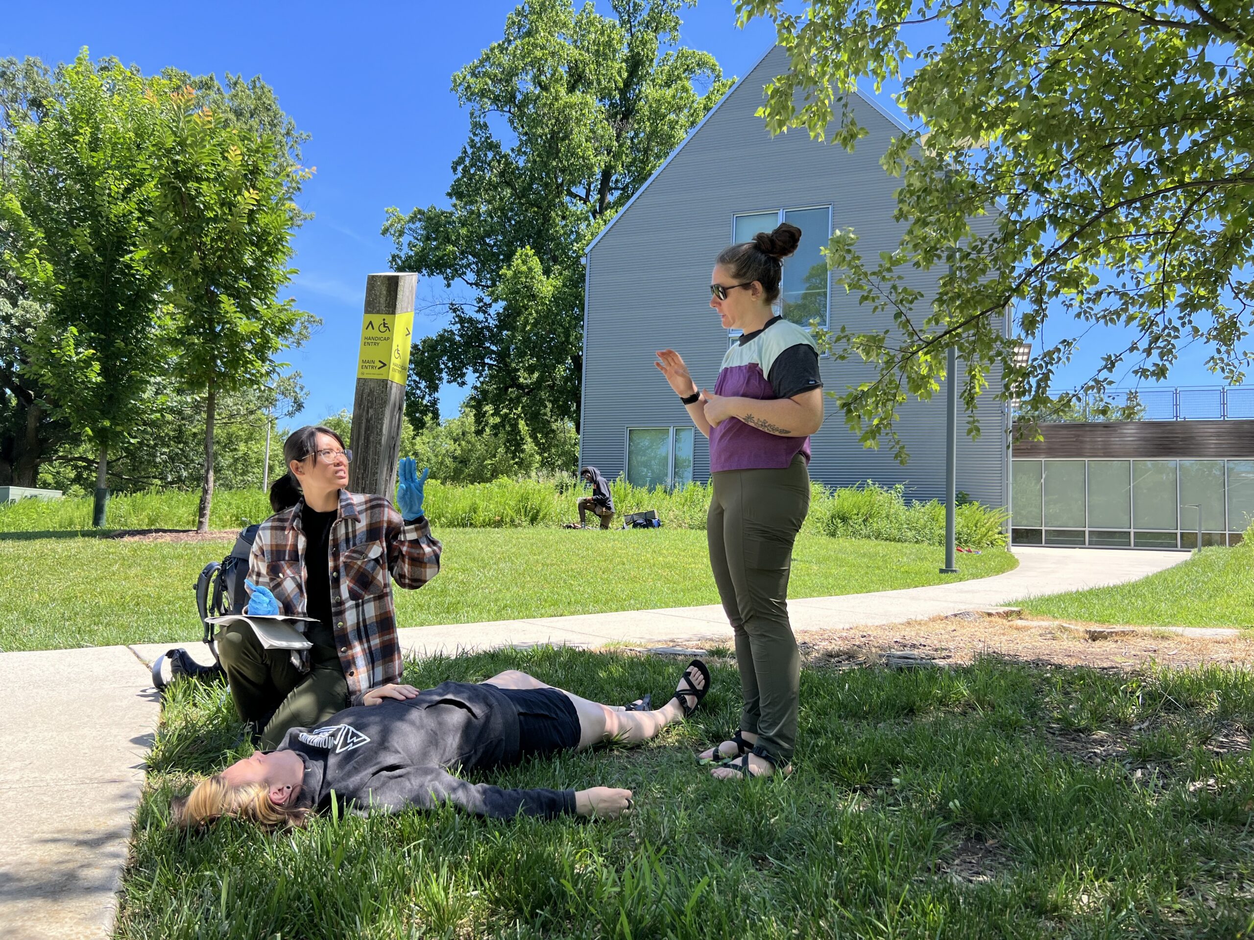 Two outdoor educators stand in a rural area. They are standing over another person. All three are in a training exercise, signing using ASL