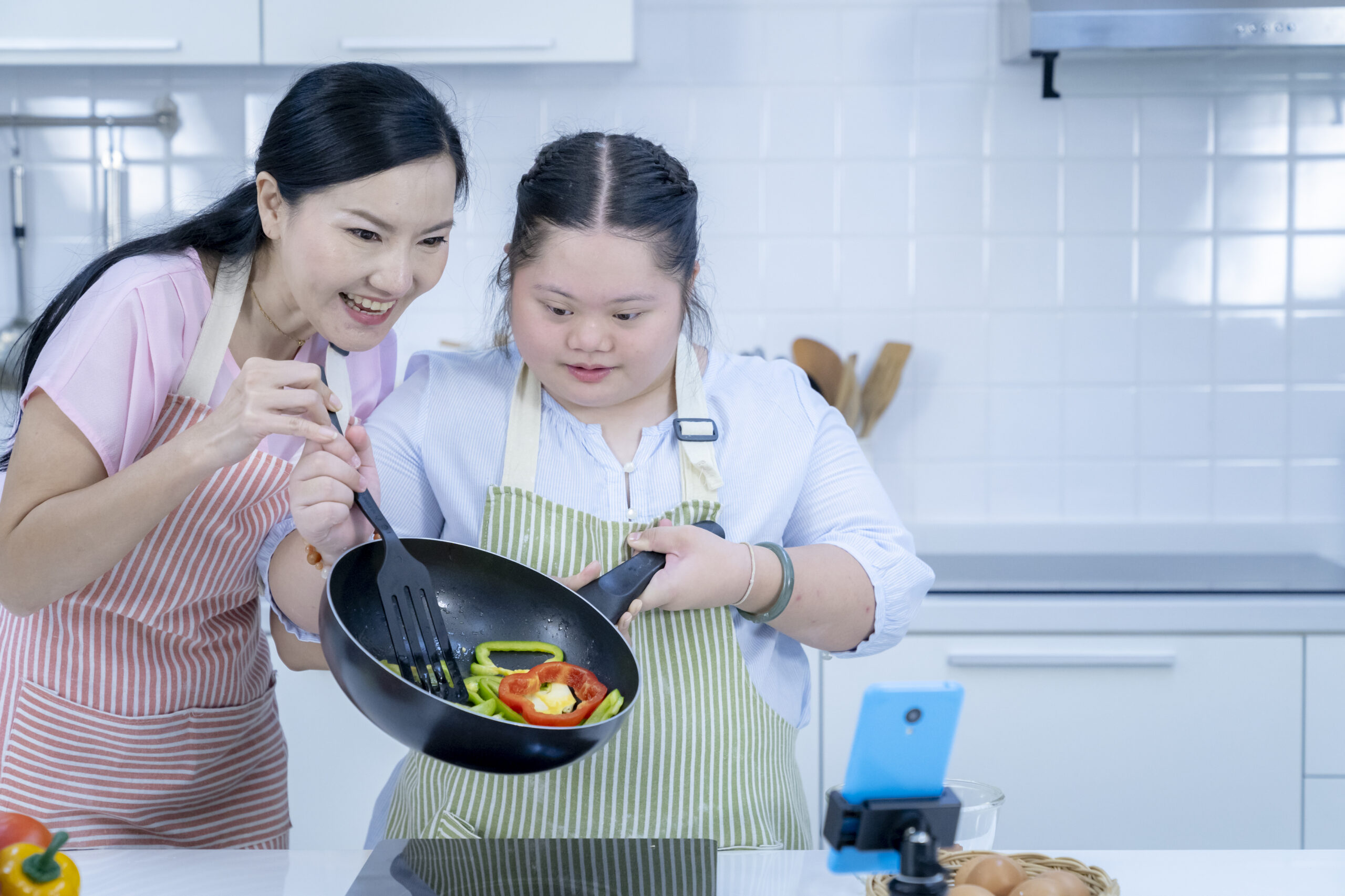 A woman and a young woman wearing aprons cook together in a bright kitchen while filming with a smartphone. The girl holds a frying pan with sliced bell peppers as the woman guides her with a spatula.