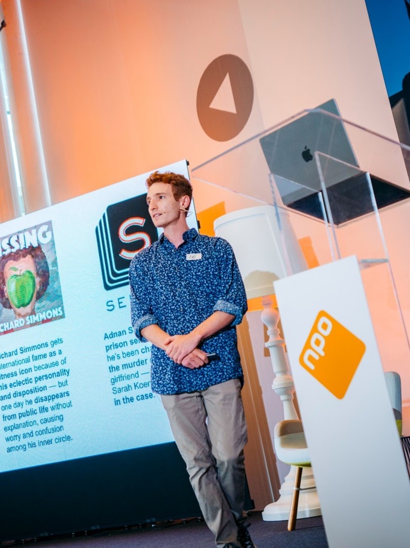 Ari stands, arms slightly crossed in front of a presentation. He is wearing a blue shirt. The background is filled with a podium, a presentation, a laptop, and a lit wall.