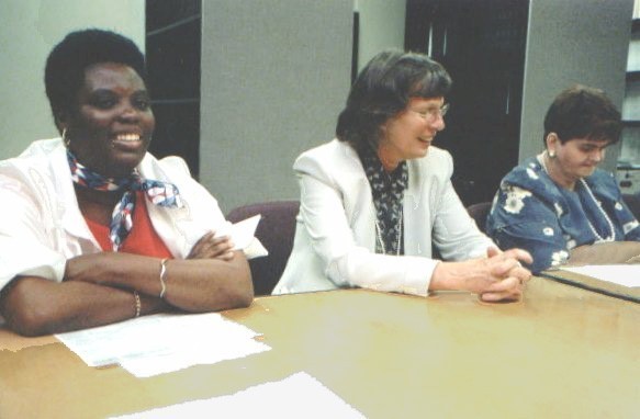 Three women sit side by side at a conference table during what appears to be a meeting. The woman on the left, wearing a white jacket and colorful scarf, is Lois Curtis, one of the lead plaintiffs in the Olmstead case. She smiles broadly with her arms folded. The woman in the center, wearing a light-colored blazer and glasses, leans forward with her hands clasped, smiling as if speaking or listening. The woman on the right, wearing a blue patterned top, looking down at papers in front of her is Elaine Wilson. Sheets of paper are spread across the table, and office shelving is visible in the background.
