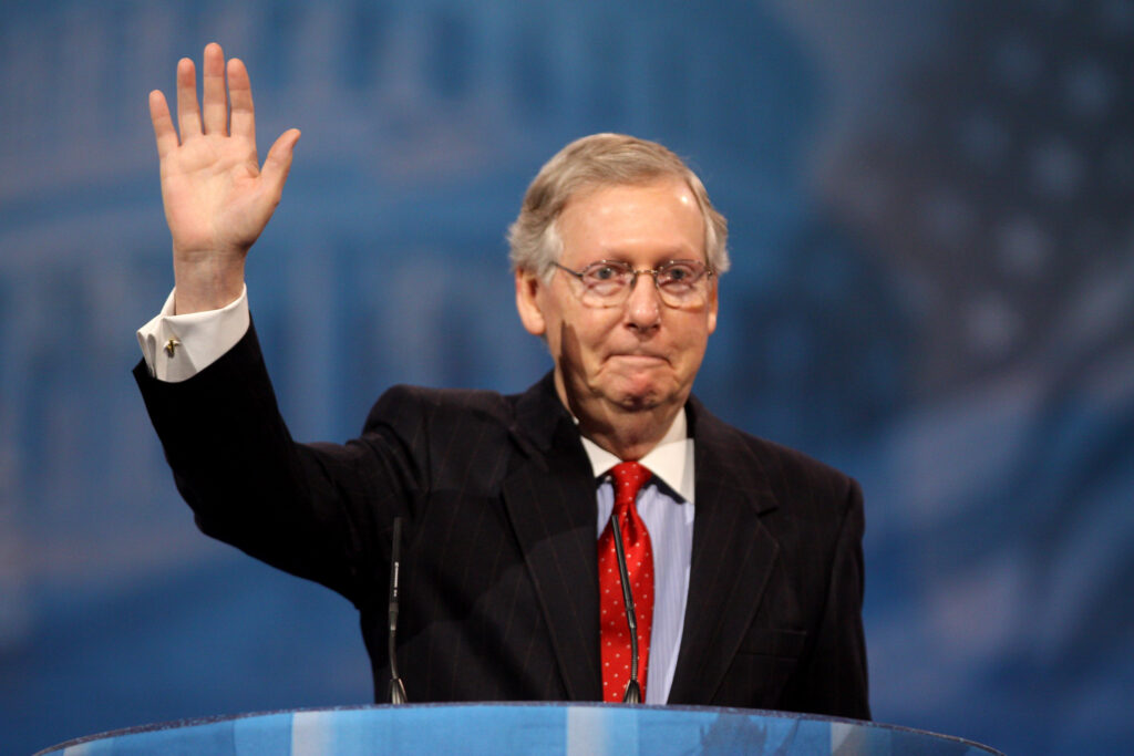Senator Mitch McConnell of Kentucky is at a podium wearing a black suit and red tie. He has his right arm raised to wave or greet attendees.
