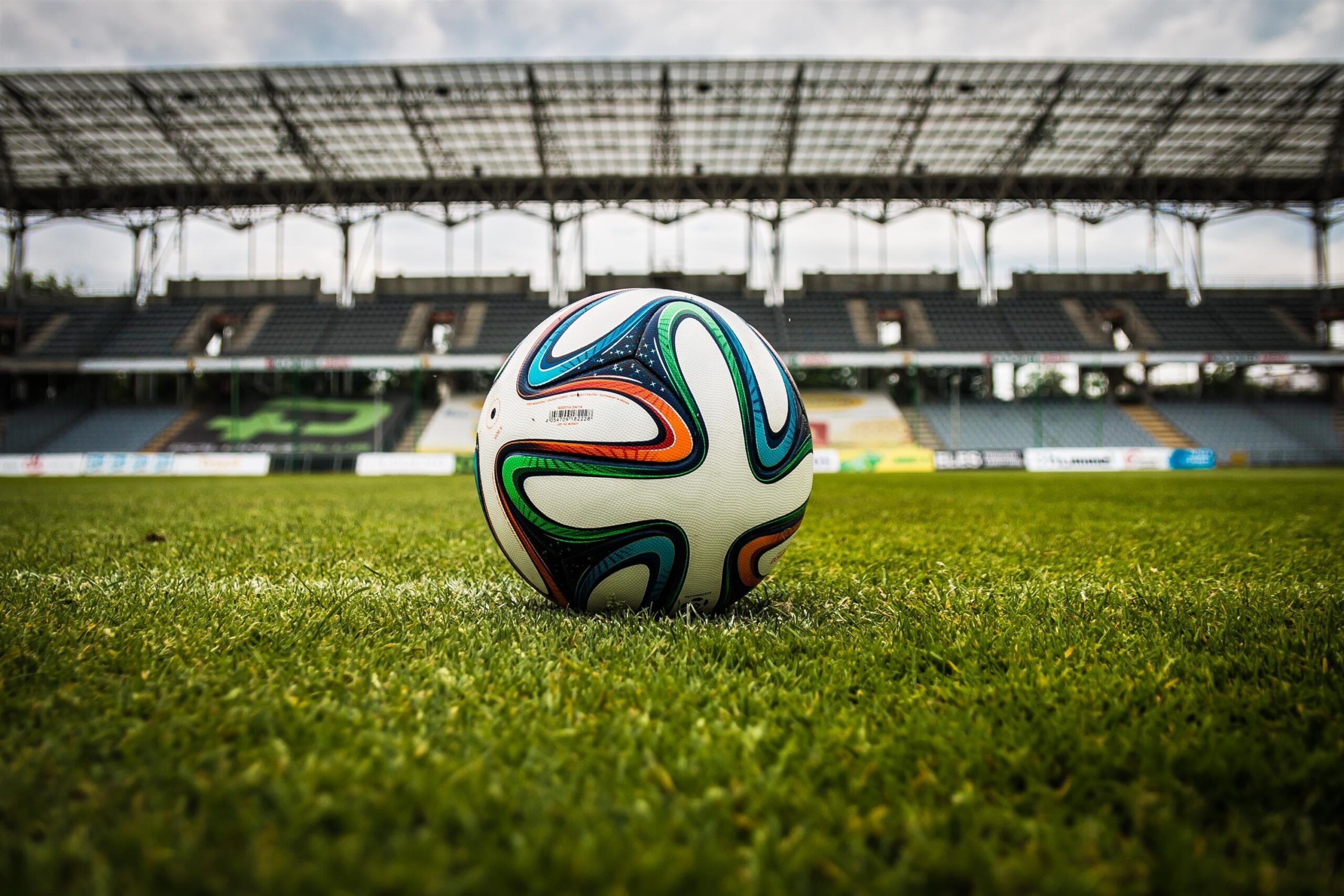 A multi-coloured soccer ball sits on the green turf of a stadium with the grandstands in the background
