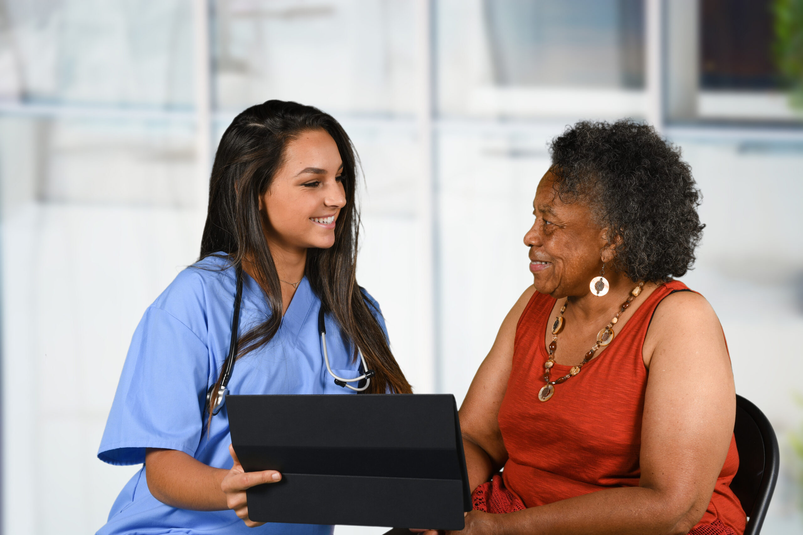 A nurse in blue scrubs smiles while showing an older woman information on a tablet in a bright healthcare setting.