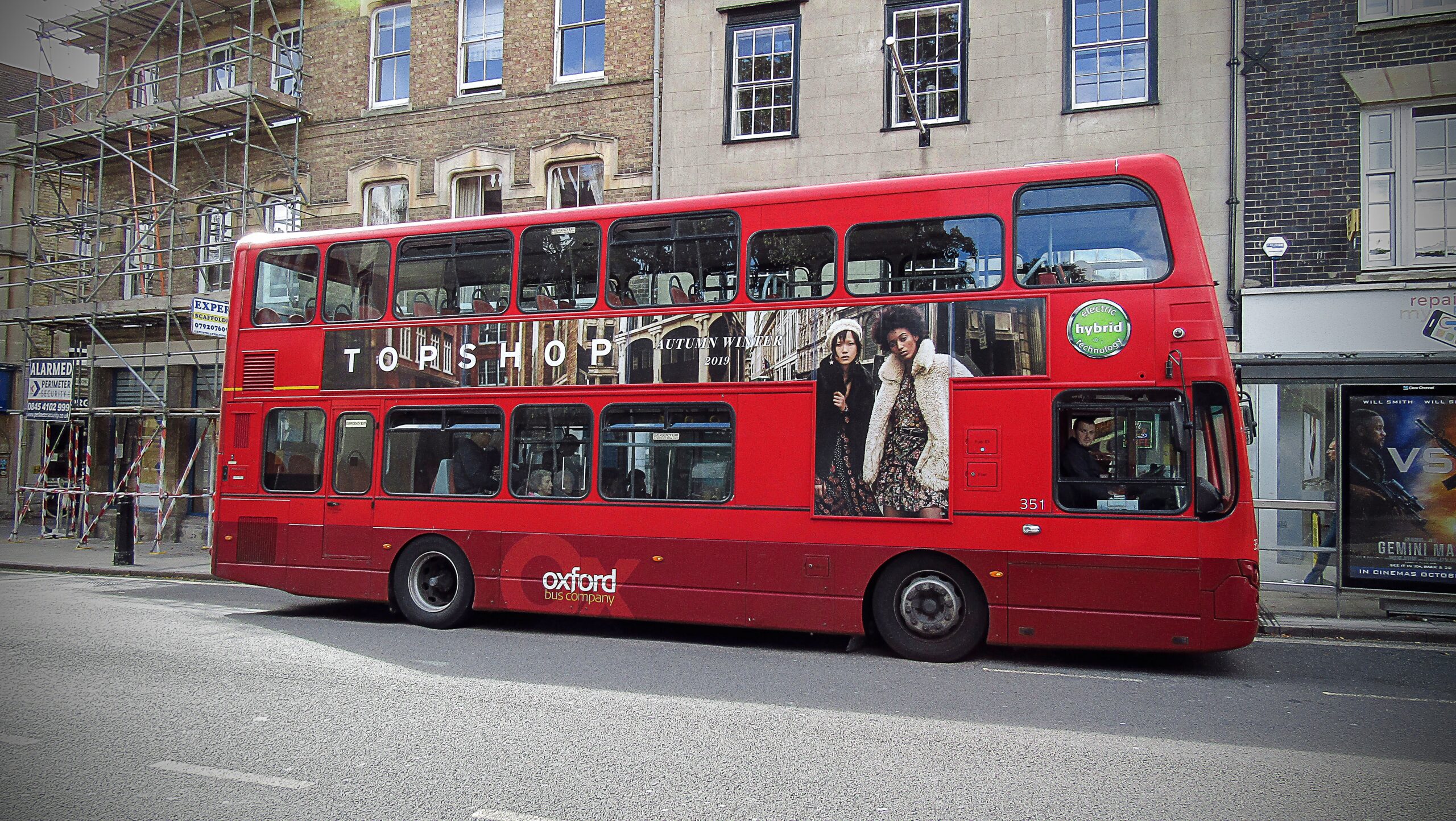 A red, British, double decker bus with an add for a high street brand is parked in front of a brick building.