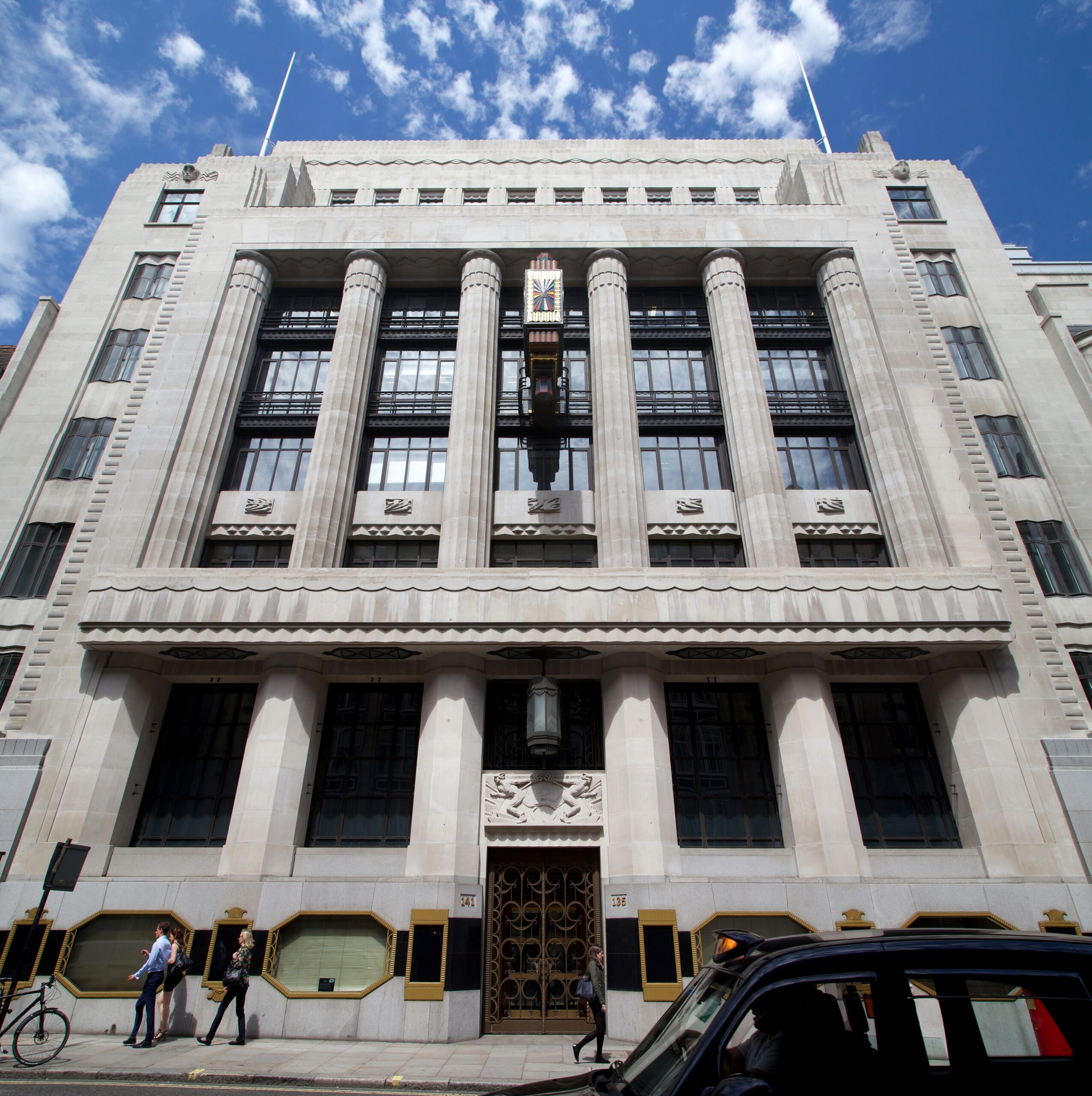 The Art Deco facade of the former Daily Telegraph Building on Fleet Street in London, a pale stone building with fluted columns, a colorful clock and gilded window frames, under a bright blue sky.