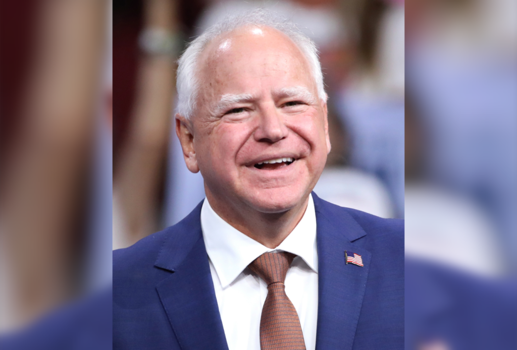 Minnesota Gov. Tim Walz smiles while wearing a navy blue suit, white shirt, rust-colored tie and American flag lapel pin. The background is blurred.