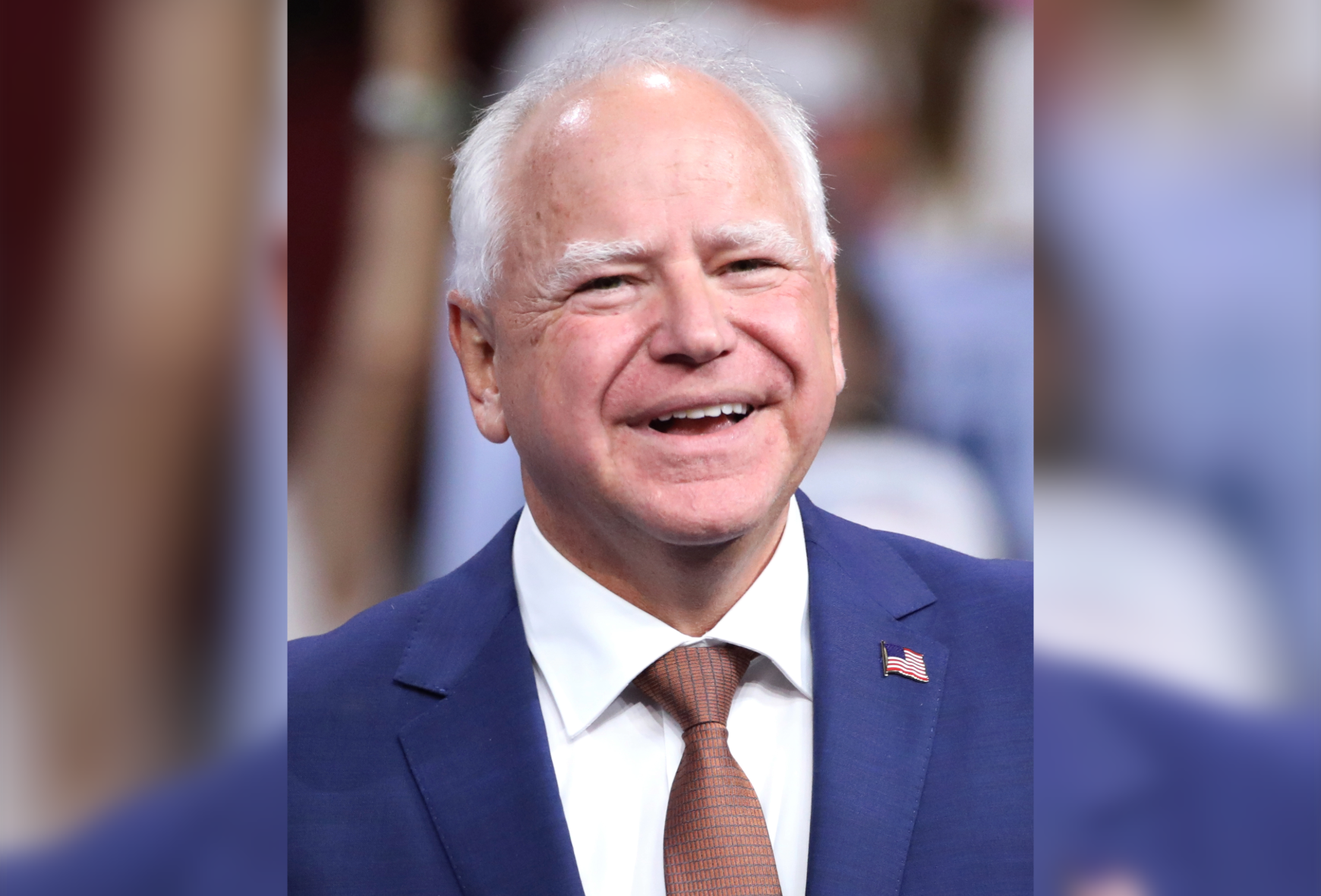 Minnesota Gov. Tim Walz smiles while wearing a navy blue suit, white shirt, rust-colored tie and American flag lapel pin. The background is blurred.